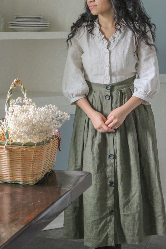 A woman in a white blouse and green skirt stands in a room with a wooden table and a basket of white flowers.